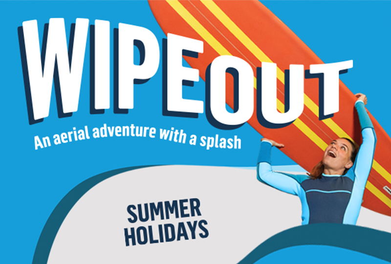 A person in a blue wetsuit lifts a bright orange surfboard against a vibrant blue backdrop, promoting summer holidays aerial adventure.